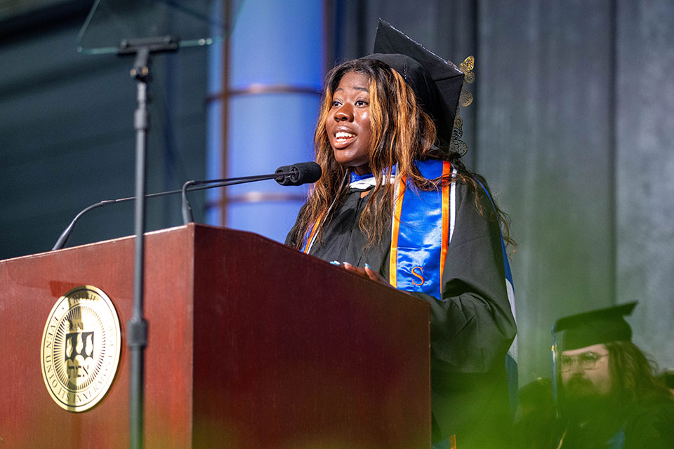 Ianna Gilbert, wearing a cap and gown, speaks from the podium