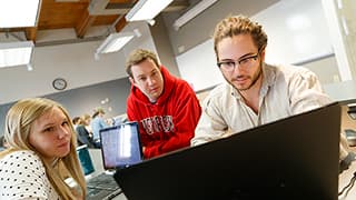 Three students gather round a laptop in a classroom.